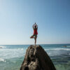 Persona ejercitando yoga sobre una roca en la playa. Envato.