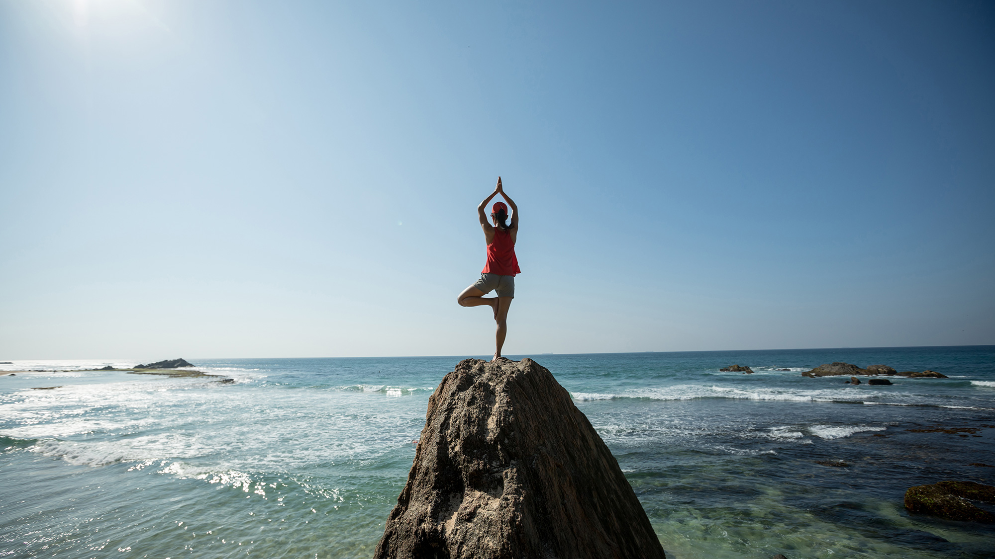 Persona ejercitando yoga sobre una roca en la playa. Envato.